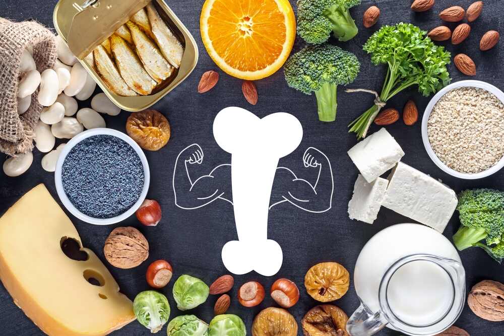 An array of calcium-rich foods placed on a chalkboard background surround a graphic of a bone with strong arms drawn in